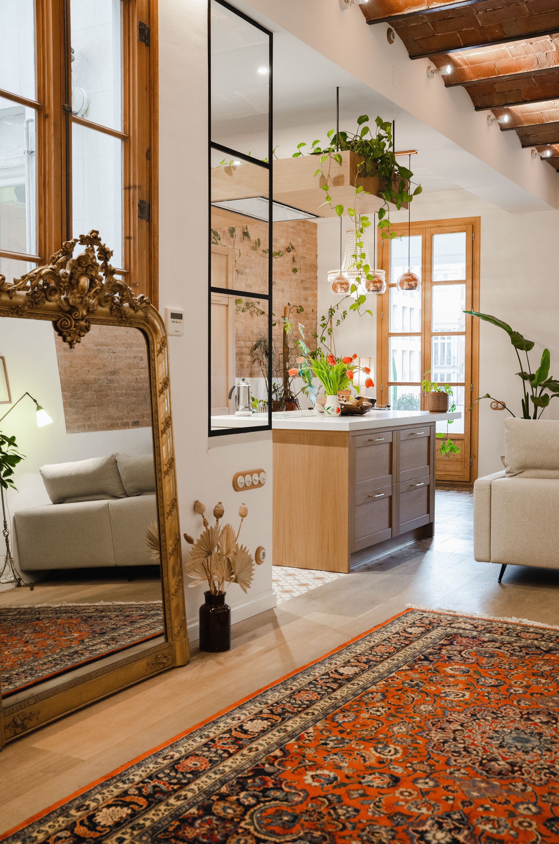 یک فضای داخلی مدرن با طراحی زیبا که ترکیبی از ظرافت و راحتی را به نمایش می‌گذارد. اتاق شامل یک آینه بزرگ و تزئینی طلایی است که گرما و هماهنگی را منعکس می‌کند. یک کاناپه خاکستری دنج به طور برجسته در فضا قرار گرفته و با یک فرش پرشین نارنجی پرجنب‌وجوش که به کفپوش چوبی روشن رنگ اضافه می‌کند، تکمیل شده است. نور طبیعی از طریق پنجره‌های بزرگ به داخل اتاق می‌تابد و منطقه‌ای پر از گیاهان را روشن می‌کند که به طراحی زندگی و شادابی می‌بخشد. فضای آشپزخانه با کابینت‌های چوبی روشن و قفسه‌های باز، حس زیبایی مدرن را ترویج می‌دهد. چراغ‌های آویز معلق نورپردازی نرم و ملایمی را فراهم می‌کنند و فضایی دلپذیر ایجاد می‌کنند. سبک کلی ترکیبی از مینیمالیسم و عناصر سنتی است که یک ترکیب زیبا و در عین حال راحت را به نمایش می‌گذارد.