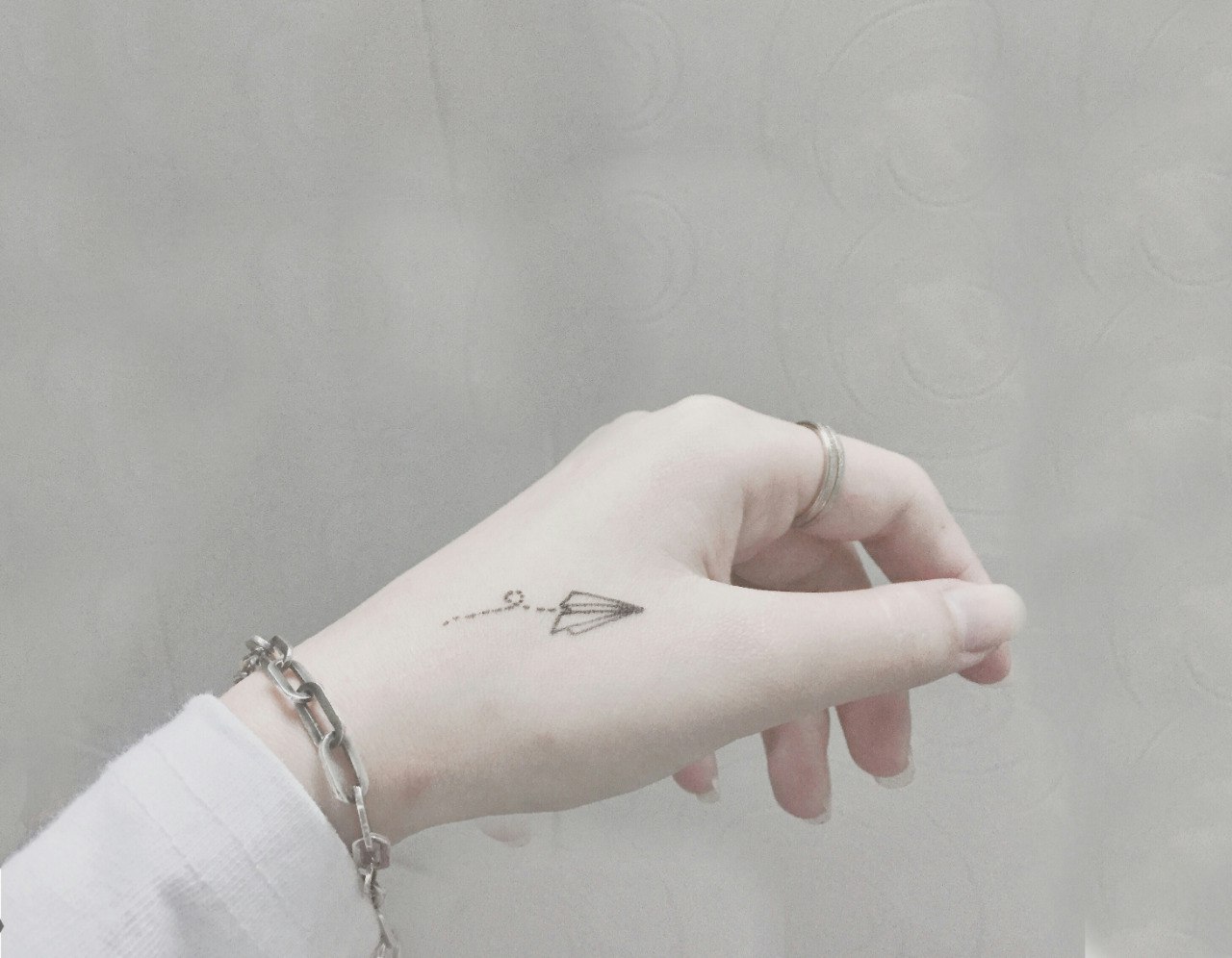 نمایی نزدیک از دستی که در برابر پس‌زمینه‌ای نرم و محو به رنگ خاکستری روشن قرار گرفته است. نقطه تمرکز یک تتوی ظریف و مینیمال از یک هواپیمای کاغذی روی مچ دست است که دارای خط‌چین‌هایی است که حرکت را نشان می‌دهد. نور طبیعی و پراکنده، تناژهای نرم پوست را تقویت کرده و جزئیات ظریف تتو را برجسته می‌کند. دست با یک دستبند نقره‌ای باریک و یک حلقه ظریف تزئین شده است که به تصویر ظرافت می‌بخشد. ترکیب‌بندی بر سادگی و لطافت تأکید دارد، با تمرکز بر تتو که مضامینی از آزادی و اکتشاف را القا می‌کند. زیبایی کلی مدرن و تمیز است و با زمینه‌های هنر یا مد معاصر هماهنگ است.